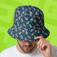 Load image into Gallery viewer, Person wearing a blue bucket hat with colorful Stumble Guys patterns on a green background
